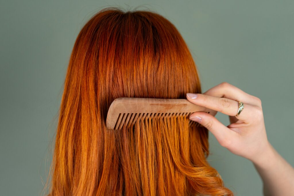 Persona peinando su cabello largo y rojizo con un peine de madera de dientes anchos, vista desde atrás. Representa cuidado capilar y prevención de la caída del cabello.