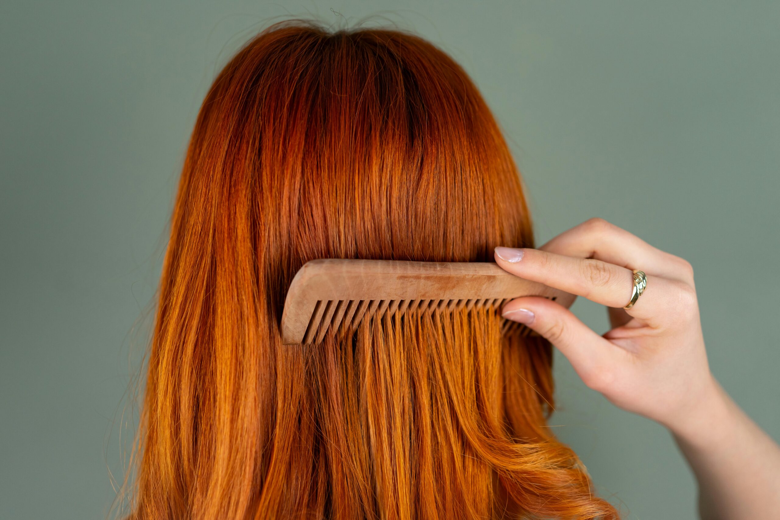 Persona peinando su cabello largo y rojizo con un peine de madera de dientes anchos, vista desde atrás. Representa cuidado capilar y prevención de la caída del cabello.